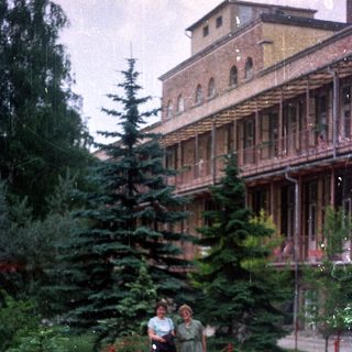 Two women stand near a building with evergreen trees.