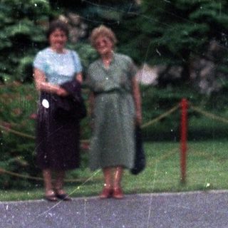 Two women stand near a building with evergreen trees.