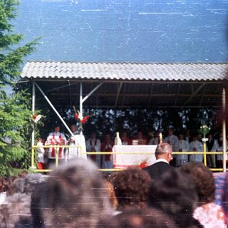 Outdoor ceremony with attendees gathered beneath a covered platform.