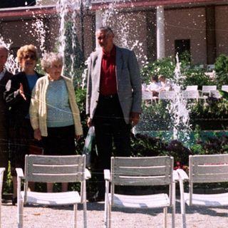 Four people stand in a garden with fountains and chairs.