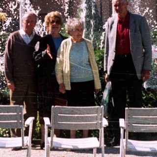 Four people stand in a garden with fountains and chairs.
