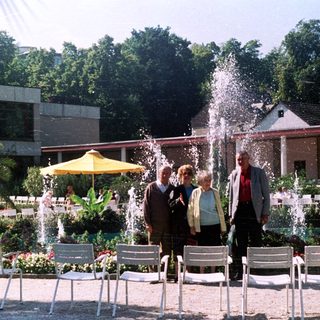Four people stand in a garden with fountains and chairs.
