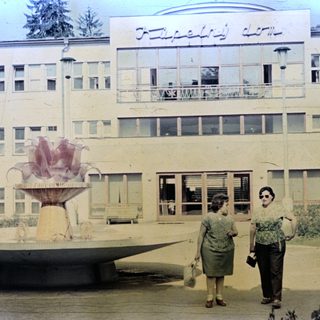 Two women stand near a fountain in front of a building.