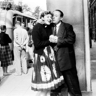 Couple embraces near a train platform, onlookers in background.