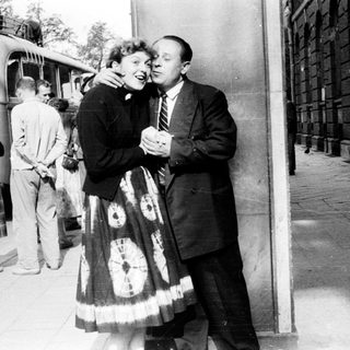 Couple embraces near a train platform, onlookers in background.