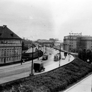 Here's a brief summary of the image: Monochrome view of a road with buildings and vehicles.