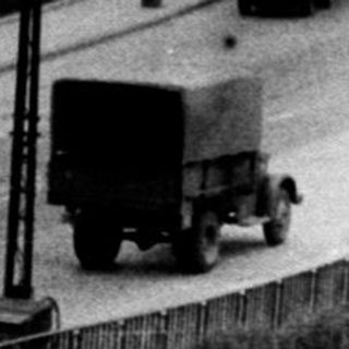 Here's a brief summary of the image: Monochrome view of a road with buildings and vehicles.