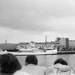 People watch boats docked near a building on a harbor.