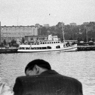 People watch boats docked near a building on a harbor.