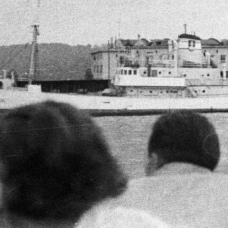 People watch boats docked near a building on a harbor.