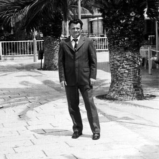 Man in suit stands near palm trees in a public space.