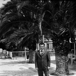Man in suit stands near palm trees in a public space.