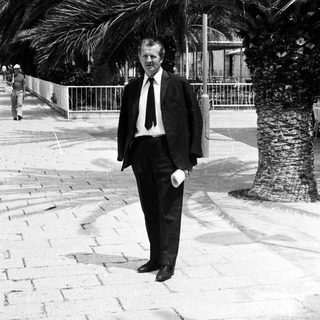 Man in suit stands near palm tree on paved street.