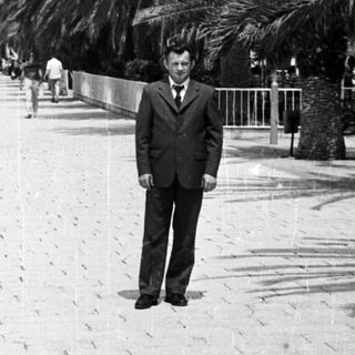Man in suit stands on a palm-lined promenade.