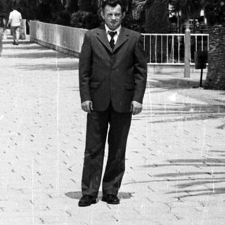 Man in suit stands on a palm-lined promenade.