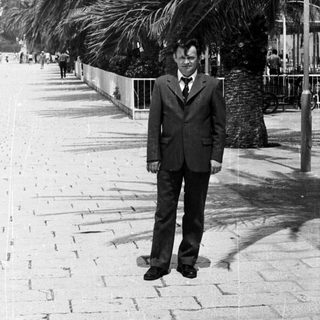 Man in suit stands on a palm-lined promenade.