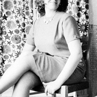 Woman seated in chair, posing near a patterned curtain.