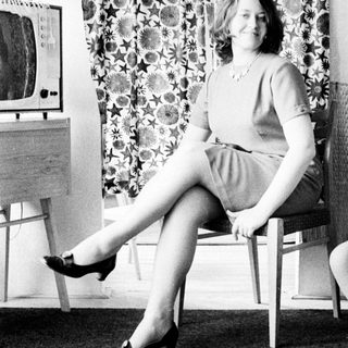 Woman seated in chair, posing near a patterned curtain.