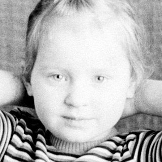 Here's a brief summary of the image: Young child seated, hands behind head, in a vintage setting.