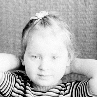 Here's a brief summary of the image: Young child seated, hands behind head, in a vintage setting.