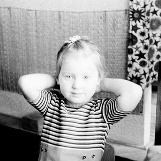 Here's a brief summary of the image: Young child seated, hands behind head, in a vintage setting.
