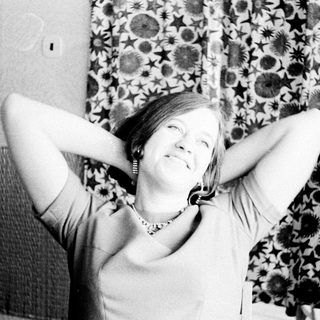 Woman smiles, arms raised behind head, patterned curtain background.