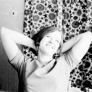 Woman smiles, arms raised behind head, patterned curtain background.