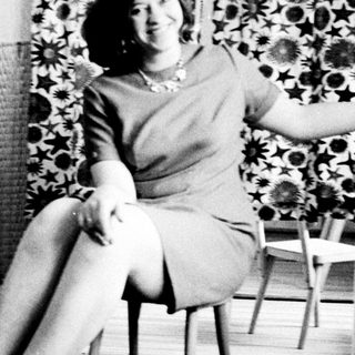 Woman in a dress sits on a chair indoors.