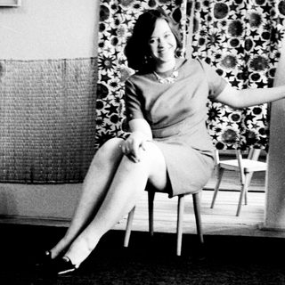 Woman in a dress sits on a chair indoors.