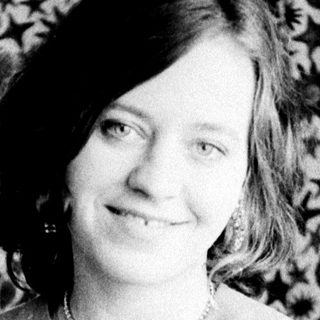 Woman in a necklace sits before a patterned wall.