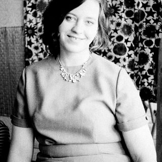 Woman in a necklace sits before a patterned wall.