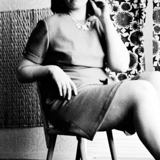 Here's a brief summary of the image: Woman in dress sits on chair, looking away.