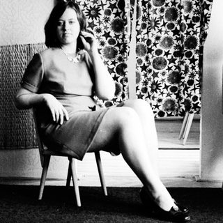 Here's a brief summary of the image: Woman in dress sits on chair, looking away.