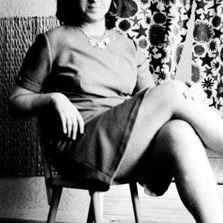 Here's a brief summary of the image: Woman seated on a chair with patterned curtains behind her.