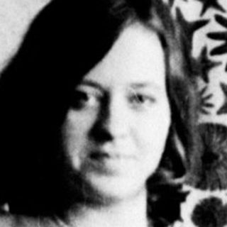 Here's a brief summary of the image: Woman seated on a chair with patterned curtains behind her.