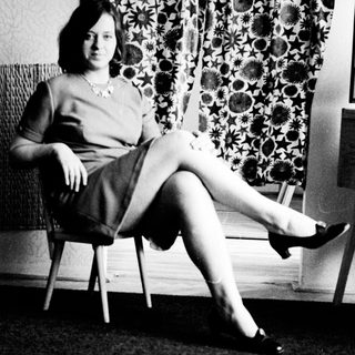 Here's a brief summary of the image: Woman seated on a chair with patterned curtains behind her.