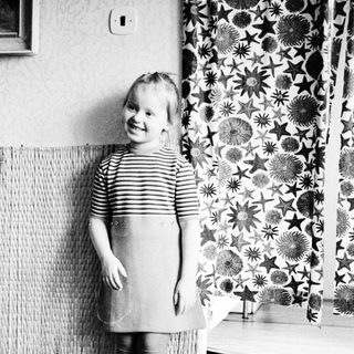 Here's a brief summary of the image: Young girl in dress stands near patterned curtains.