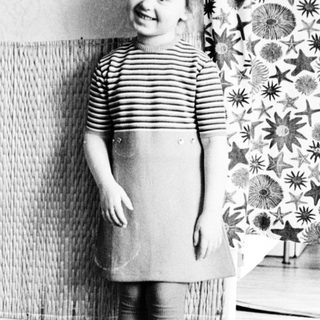 Here's a brief summary of the image: Young girl in dress stands near patterned curtains.