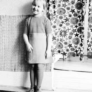 Here's a brief summary of the image: Young girl in dress stands near patterned curtains.