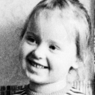 Here's a brief summary of the image: Young girl in dress stands near patterned curtains.