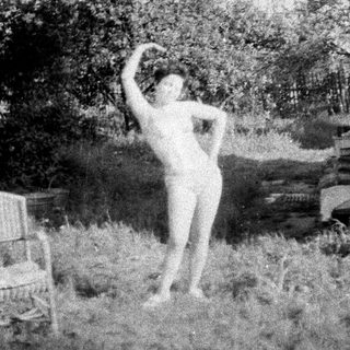 Here's a brief summary of the image: Nude woman stands outdoors, arms raised, near a building.