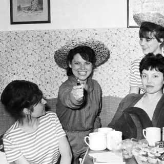 Here's a brief summary of the image: Women seated indoors, enjoying refreshments, wearing hats.