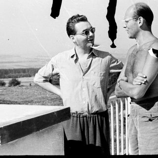 Two men converse on a balcony with a view.