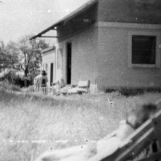 Here's a brief summary of the image: Old house with overgrown grass and a person on the porch.