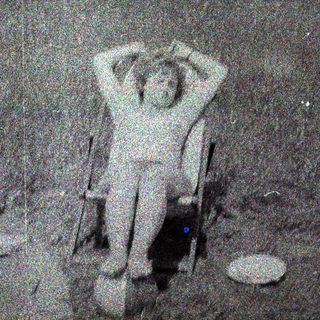 Person seated on chair, arms raised, in an outdoor setting.