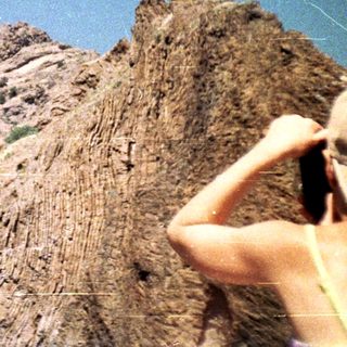 A person photographs a rocky mountain landscape under a blue sky.