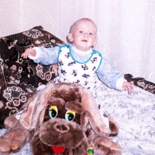 Baby sits with a large plush dog on a bed.