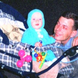 Man pushes stroller with baby at dusk near building.