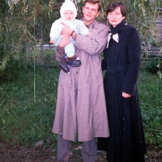 Man, woman, and baby stand outdoors near a tree.