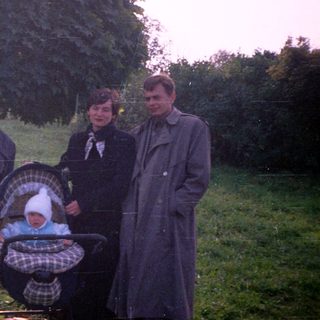 Family stands near a stroller in a grassy, wooded area.